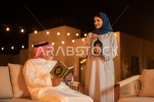 التقرب إلى الله في شهر رمضان المبارك, تلاوة وحفظ المصحف الشريف، رجل كبير بالسن عربي سعودي خليجي يرتدي الشماغ والثوب التقليدي يحمل بيده القرآن الكريم، الجلوس في الهواء الطلق في حديقة إحدى الشاليهات الفاخرة، امرأة محجبة تحمل بيدها فانوس معدني مزخرف مضيء