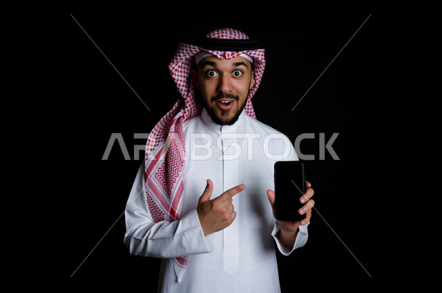 بورتريه لشاب خليجي عربي يشير إلى الهاتف الخلوي بدهشة , خلفية سوداء 