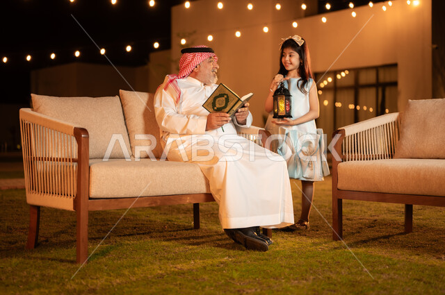 تربية وتعليم الأبناء على العبادة والتقرب من الله تعالى, تلاوة وحفظ المصحف الشريف في شهر رمضان المبارك, فوانيس وزينة الاعياد والمناسبات الاسلامية, رجل كبير بالسن عربي خليجي سعودي يرتدي الزي التقليدي يحمل بيده القرآن الكريم ويجلس في حديقة المنزل برفقة حفيدته