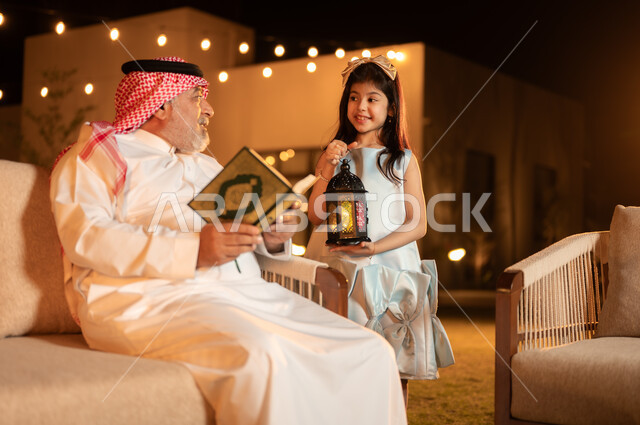  تلاوة وحفظ المصحف الشريف في شهر رمضان المبارك, تربية وتعليم الأبناء على العبادة والتقرب من الله تعالى, فوانيس وزينة الاعياد والمناسبات الاسلامية, رجل كبير بالسن عربي خليجي سعودي يرتدي الزي التقليدي يحمل بيده القرآن الكريم ويجلس في حديقة المنزل برفقة حفيدته