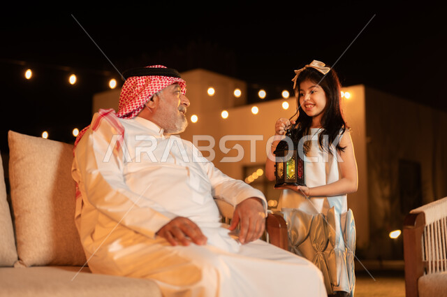 أجواء المجتمع السعودي في الأعياد والمناسبات الاسلامية, الاحتفال مع الأطفال بقدوم شهر رمضان المبارك, فتاة عربية خليجية سعودية ترتدي الفستان تحمل بيدها فانوس مضيء برفقة جدها, مفهوم احترام كبار السن, لحظات عائلية مليئة بالحب