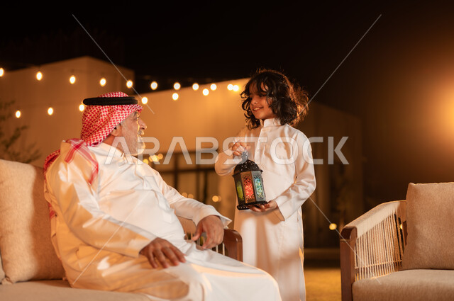 فرحة الأطفال بقدوم شهر الخير, مفهوم احترام كبار السن, فتى عربي خليجي سعودي يرتدي الثوب التقليدي يمسك بيده فانوس متوهج ويقضي وقت ممتع برفقة جده في حديقة المنزل, لحظات وأجواء مليئة بالحب والحنان, زينة وإضاءة الأعياد والمناسبات السعيدة