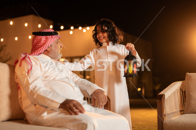 مفهوم احترام كبار السن, فتى عربي خليجي سعودي يرتدي الثوب التقليدي يمسك بيده فانوس متوهج ويقضي وقت ممتع برفقة جده في حديقة المنزل, لحظات وأجواء مليئة بالحب والحنان, زينة وإضاءة الأعياد والمناسبات السعيدة, فرحة الأطفال بقدوم شهر الخير