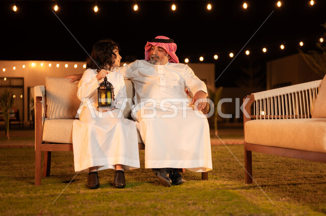 نظرات وجلسات مليئة بالحب والحنان واللطافة, زينة وإضاءة الأعياد والمناسبات السعيدة, فرحة الأطفال بقدوم شهر الخير, مفهوم احترام كبار السن, فتى عربي خليجي سعودي يرتدي الثوب التقليدي يمسك بيده فانوس متوهج ويجلس برفقة جده في حديقة المنزل