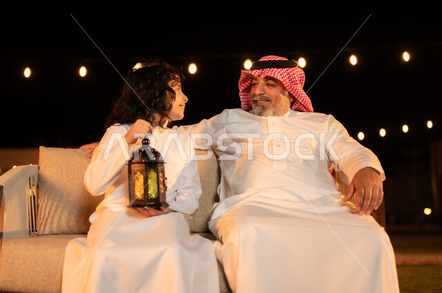 نظرات وجلسات مليئة بالحب والحنان واللطافة, زينة وإضاءة الأعياد والمناسبات السعيدة, فرحة الأطفال بقدوم شهر الخير, مفهوم احترام كبار السن, فتى عربي خليجي سعودي يرتدي الثوب التقليدي يمسك بيده فانوس متوهج ويجلس برفقة جده في حديقة المنزل