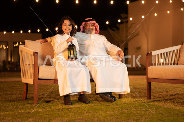 Decorations and lighting for holidays and happy occasions, children’s joy at the arrival of the month of goodness, the concept of respecting the elderly, a Saudi Gulf Arab boy wearing a traditional dress holding a glowing lantern in his hand and sitting with his grandfather in the garden of the house, moments and atmospheres full of love and tenderness