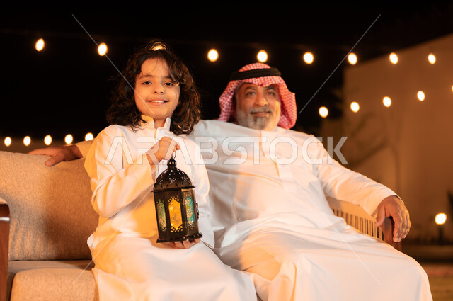 زينة وإضاءة الأعياد والمناسبات السعيدة, فرحة الأطفال بقدوم شهر الخير, مفهوم احترام كبار السن, فتى عربي خليجي سعودي يرتدي الثوب التقليدي يمسك بيده فانوس متوهج ويجلس برفقة جده في حديقة المنزل, لحظات وأجواء مليئة بالحب والحنان 