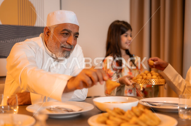 ايماءات السرور لاجتماع العائلة حول مائدة إفطار رمضان المبارك، موعد كسر الصيام عند أذان المغرب، صورة مقربة لرجل كبير بالسن عربي خليجي سعودي مبتسم يرتدي الثوب التقليدي يجلس مع اسرته حول سفرة شهر الخير، أجواء عائلية روحانية دينية إسلامية