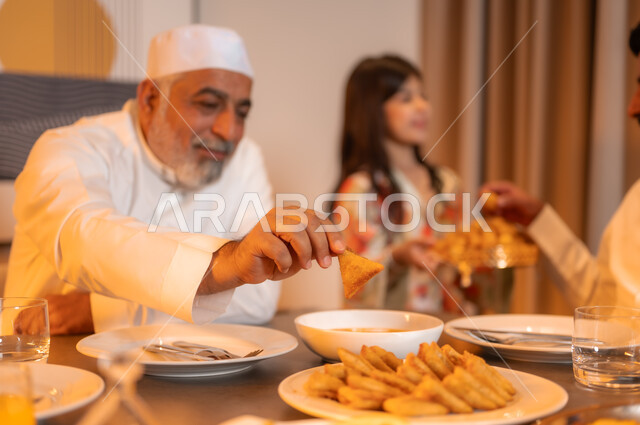 ايماءات السرور لاجتماع العائلة حول مائدة إفطار رمضان المبارك، موعد كسر الصيام عند أذان المغرب، صورة مقربة لرجل كبير بالسن عربي خليجي سعودي مبتسم يرتدي الثوب التقليدي يجلس مع اسرته حول سفرة شهر الخير، أجواء عائلية روحانية دينية إسلامية