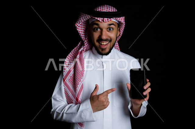 بورتريه لشاب خليجي عربي يشير إلى الهاتف الخلوي بدهشة , خلفية سوداء 