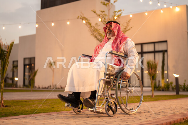 الاستمتاع بالتجول في حديقة المنزل, دعم المجتمع السعودي لذوي الاحتياجات الخاصه, شعور ذوي الهمم بالراحه والطمأنينه في بيئتهم, رجل كبير بالسن عربي خليجي سعودي مقعد يرتدي الثوب التقليدي  والشماغ يجلس على كرسي متحرك في إحدى الشاليهات في المملكة العربية السعودية