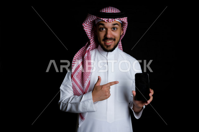 بورتريه لشاب خليجي عربي يشير إلى الهاتف الخلوي بدهشة , خلفية سوداء 