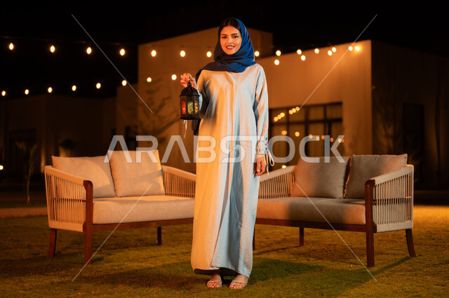 قضاء وقت ممتع في إحدى الشاليهات الفاخرة، الأجواء الرمضانية الروحانية، إضاء وزينة المناسبات الدينية الإسلامية في شهر رمضان المبارك, امرأة عربية خليجية سعودية محجبة ترتدي العباءة تحمل بيدها فانوس معدني مضيء تنظر إلى الكاميرار بتعابير السرور