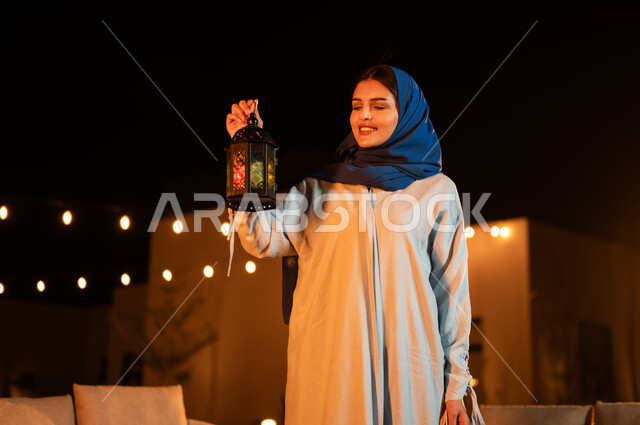 الأجواء الرمضانية الروحانية، قضاء وقت ممتع في إحدى الشاليهات الفاخرة، إضاء وزينة المناسبات الدينية الإسلامية في شهر رمضان المبارك, امرأة عربية خليجية سعودية محجبة ترتدي العباءة تحمل بيدها فانوس معدني مزخرف مضيء 