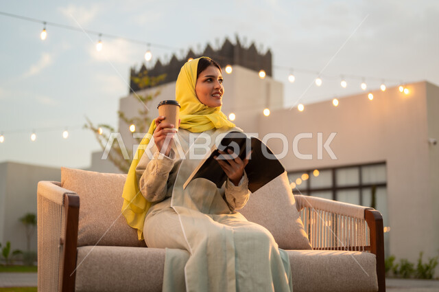 الاستمتاع بالمشروبات الساخنة في الخارج, مفهوم الثقافة والاطلاع والتعلم الذاتي, إمرأة عربية خليجية سعودية ترتدي العباءة والحجاب الأصفر تقوم بالقراءة والمطالعة في الهواء الطلق, الشغف بممارسة الهوايات المفيدة, الأنشطة الترفيهية الممتعه