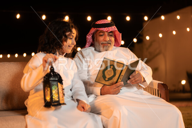 فوانيس وزينة الاعياد والمناسبات الاسلامية, تلاوة وحفظ المصحف الشريف في شهر رمضان المبارك, تربية وتعليم الأبناء على العبادة والتقرب من الله تعالى, رجل كبير بالسن عربي خليجي سعودي يرتدي الزي التقليدي يحمل بيده القرآن الكريم ويجلس في حديقة المنزل برفقة حفيده