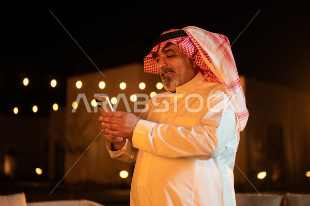 دردشه ومحادثات مع الاصدقاء والاقارب، قضاء أوقات ممتعة في إحدى حدائق الشاليهات الفاخرة في السعودية، نزهه في الهواء الطلق، استخدام الاجهزة التقنية الحديثة, صورة مقربة لرجل عربي خليجي سعودي كبير بالسن يرتدي الشماغ والثوب التقليدي يستخدم الهاتف المحمول