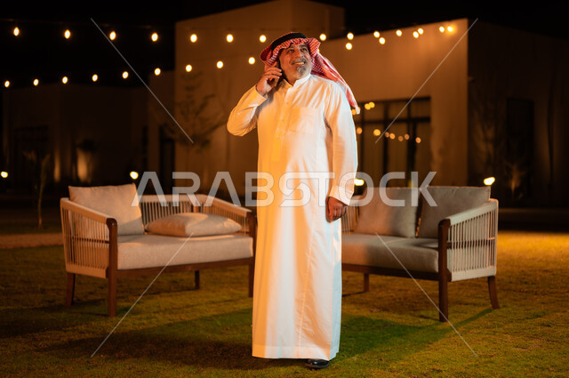إجراء مكالمه صوتية عبر الهاتف المحمول، نزهه في الهواء الطلق، استخدام الاجهزة التقنية الحديثة, رجل عربي خليجي سعودي كبير بالسن يرتدي الشماغ والثوب التقليدي وقضي أوقات ممتعة في إحدى حدائق الشاليهات الفاخرة في السعودية، دردشه ومحادثات مع الاصدقاء والاقارب