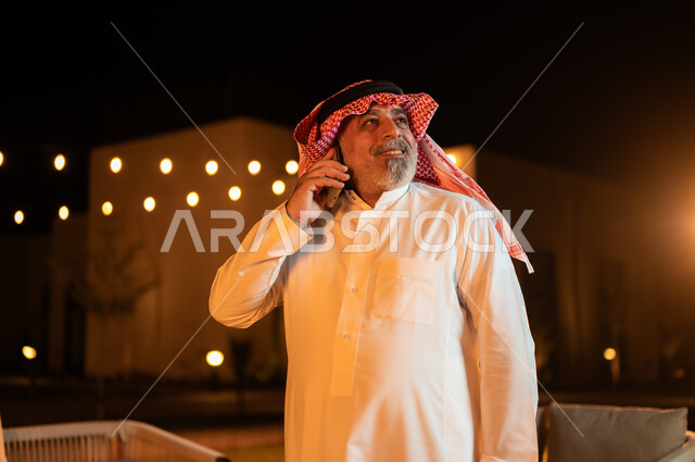 نزهه في الهواء الطلق، استخدام الاجهزة التقنية الحديثة, إجراء مكالمه صوتية عبر الهاتف المحمول، صورة مقربة لرجل عربي خليجي سعودي كبير بالسن يرتدي الشماغ والثوب التقليدي وقضي أوقات ممتعة في إحدى حدائق الشاليهات الفاخرة في السعودية، دردشه ومحادثات مع الاصدقاء والاقارب