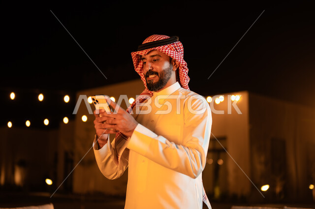 دردشه ومحادثات مع الاصدقاء والاقارب، قضاء أوقات ممتعة في إحدى حدائق الشاليهات الفاخرة في السعودية، نزهه في الهواء الطلق، استخدام الاجهزة التقنية الحديثة, صورة مقربة لرجل عربي خليجي سعودي يرتدي الشماغ والثوب التقليدي يستخدم الهاتف المحمول