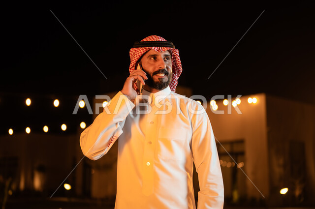 نزهه في الهواء الطلق، استخدام الاجهزة التقنية الحديثة, إجراء مكالمه صوتية عبر الهاتف المحمول، صورة مقربة لرجل عربي خليجي سعودي يرتدي الشماغ والثوب التقليدي وقضي أوقات ممتعة في إحدى حدائق الشاليهات الفاخرة في السعودية، دردشه ومحادثات مع الاصدقاء والاقارب