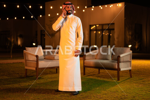 استخدام الاجهزة التقنية الحديثة, إجراء مكالمه صوتية عبر الهاتف المحمول، رجل عربي خليجي سعودي يرتدي الشماغ والثوب التقليدي وقضي أوقات ممتعة في إحدى حدائق الشاليهات الفاخرة في السعودية، دردشه ومحادثات مع الاصدقاء والاقارب, نزهه في الهواء الطلق