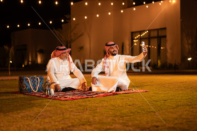 توثيق اللحظات السعيدة، مفهوم حسن الضيافه والاستقبال، رجل عربي خليجي سعودي يقوم بالتقاط صور السيلفي التذكارية مع والده باستخدام الهاتف الجوال، قضاء أوقات ممتعة في إحدى حدائق الشاليهات الفاخرة في السعودية، نزهة في الهواء الطلق