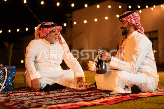 Hanging out and exchanging conversations in an outdoor session, the concept of good hospitality and reception, pouring coffee from the copper pot, an old Saudi Gulf Arab man wearing a shemagh and traditional dress sitting with his son on the grass, spending enjoyable times in one of the gardens of the luxurious chalets in Saudi Arabia.
