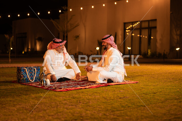 مفهوم حسن الضيافه والاستقبال، التسامر وتبادل الحديث في جلسة خارجية، رجل عربي خليجي سعودي كبير بالسن يرتدي الشماغ والثوب التقليدي يجلس مع ابنه على العشب ويقومان بتبادل الأحاديث، قضاء أوقات ممتعة في إحدى حدائق الشاليهات الفاخرة في السعودية
