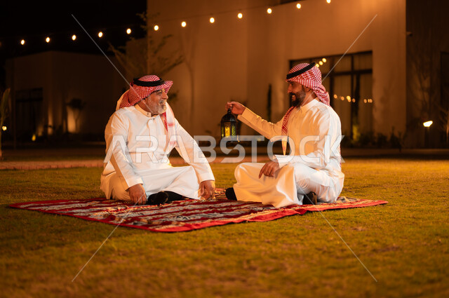 زينة واكسسوارات شهر رمضان المبارك، التسامر وتبادل الحديث في جلسة خارجية، رجل عربي خليجي سعودي يرتدي الشماغ والثوب التقليدي يحمل فانوس معدني مضيء يجلس مع والده على العشب ويقومان بتبادل الأحاديث، قضاء أوقات ممتعة في إحدى حدائق الشاليهات الفاخرة في السعودية