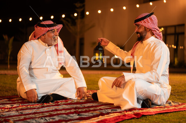 زينة واكسسوارات شهر رمضان المبارك، التسامر وتبادل الحديث في جلسة خارجية، رجل عربي خليجي سعودي يرتدي الشماغ والثوب التقليدي يحمل فانوس معدني مضيء يجلس مع والده على العشب ويقومان بتبادل الأحاديث، قضاء أوقات ممتعة في إحدى حدائق الشاليهات الفاخرة في السعودية