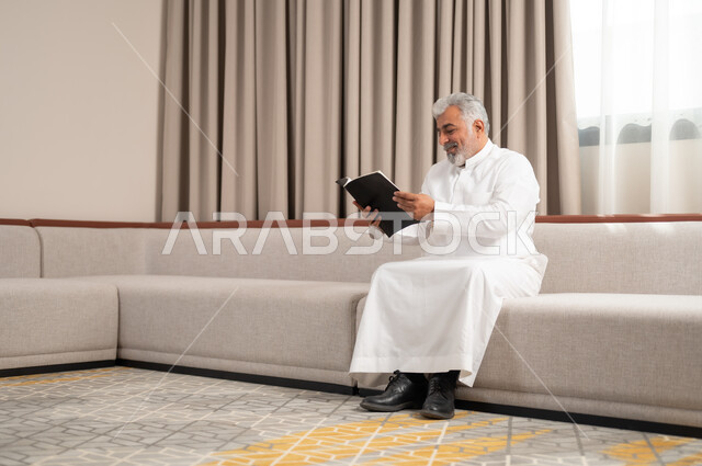 الاستمتاع بممارسة هواية المطالعة، رجل كبير السن عربي خليجي سعودي مبتسم يرتدي الزي التقليدي يجلس على كنبة مريحة يقوم بقراءة كتاب بإيماءات السعادة والسرور، مفهوم التثقيف والمعرفة، القصص والروايات الممتعة