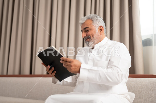 الاستمتاع بممارسة هواية المطالعة، صورة مقربة لرجل كبير السن عربي خليجي سعودي مبتسم يرتدي الزي التقليدي يجلس على كنبة مريحة يقوم بقراءة كتاب بإيماءات السعادة والسرور، مفهوم التثقيف والمعرفة، القصص والروايات الممتعة