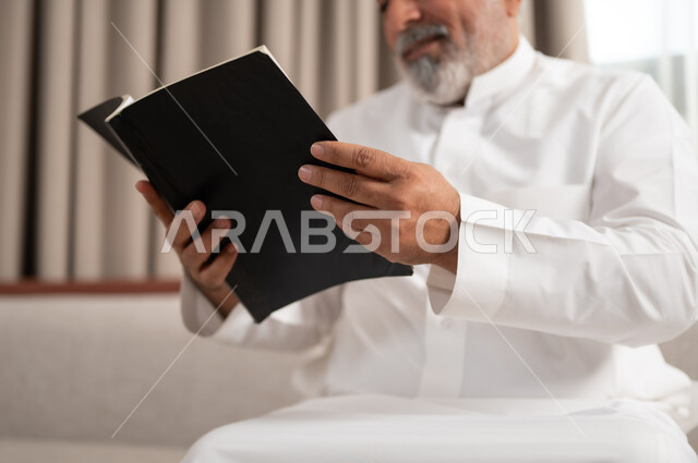 الاستمتاع بممارسة هواية المطالعة، صورة مقربة لرجل كبير السن عربي خليجي سعودي مبتسم يرتدي الزي التقليدي يجلس على كنبة مريحة يقوم بقراءة كتاب بإيماءات السعادة والسرور، مفهوم التثقيف والمعرفة، القصص والروايات الممتعة