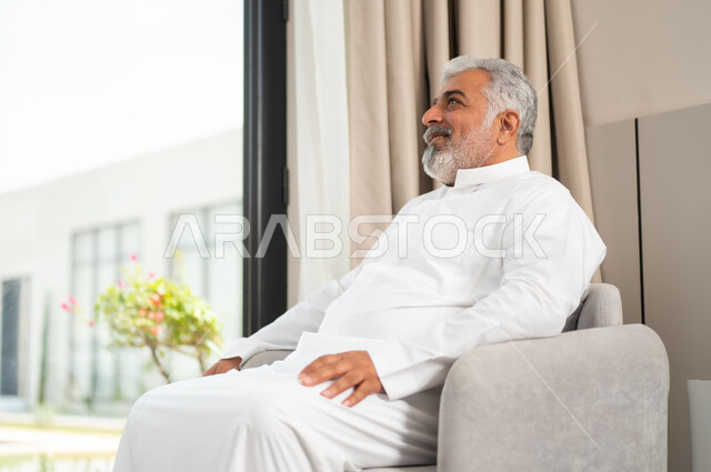 الاستمتاع بمشاهدة التلفاز، مفهوم التحديق والتركيز، صورة مقربة لرجل كبير السن عربي خليجي سعودي مبتسم يرتدي الزي التقليدي يجلس على كنبة مريحة ينظر إلى شيء ما بإيماءات التأمل، متابعة الأفلام والبرامج المفضلة