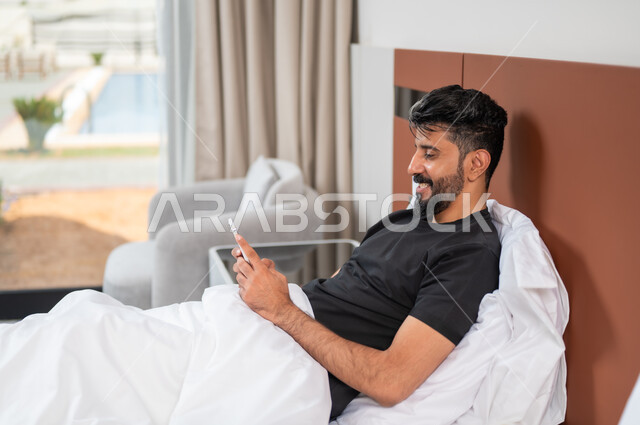 الدردشة والتواصل مع الاصدقاء بإيماءات السعادة والفرح، استخدام الأجهزة والتقنيات الحديثة، صورة مقرب لشاب عربي خليجي سعودي مبتسم يجلس على السرير يمسك بيده الهاتف المحمول يقوم بتصفح الانترنت ومواقع التواصل الاجتماعي
