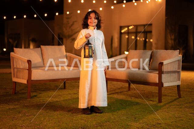 Preparing to welcome the month of goodness, happy customs and traditions in the holy month of Ramadan, lighting and decorations for holidays and occasions in the Kingdom of Saudi Arabia, a smiling Saudi Gulf Arab boy wearing a traditional dress holding a luminous metal lantern in his hand and looking at the camera