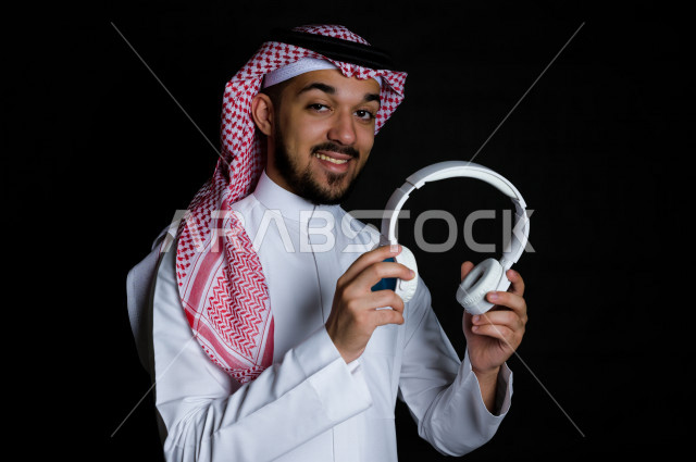بورتريه لشاب سعودي خليجي عربي يمسك بيده سماعة لاسلكية بيضاء , خلفية سوداء 