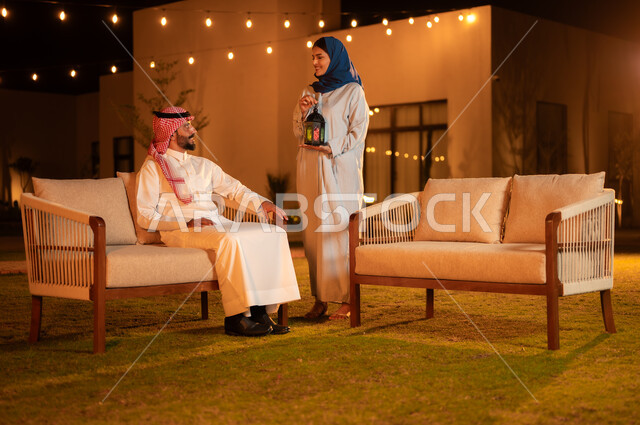 تبادل نظرات الحب والمودة بين الزوجين، إمرأة عربية خليجية سعودية مبتسمة ترتدي الحجاب تحمل فانوس معدني مضيء تقف بجانب زوجها في الجلسة الخارجية لاحد الشاليهات الراقية، الأجواء الرمضانية العائلية الروحانية، زينة واكسسوارات شهر رمضان المبارك
