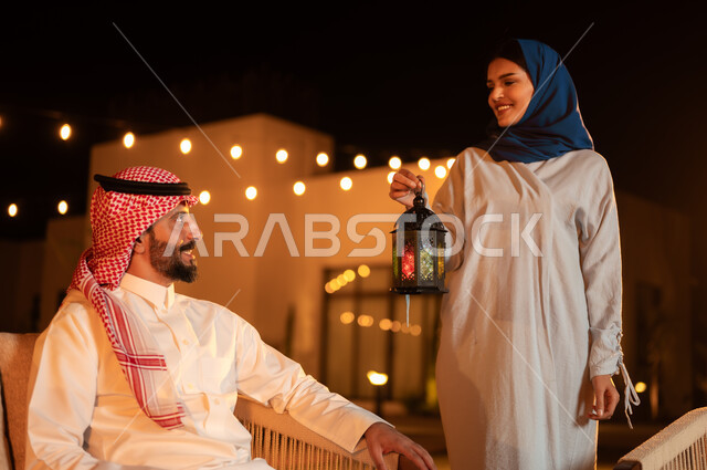 تبادل نظرات الحب والمودة بين الزوجين، إمرأة عربية خليجية سعودية مبتسمة ترتدي الحجاب تحمل فانوس معدني مضيء تقف بجانب زوجها في الجلسة الخارجية لاحد الشاليهات الراقية، الأجواء الرمضانية العائلية الروحانية، زينة واكسسوارات شهر رمضان المبارك