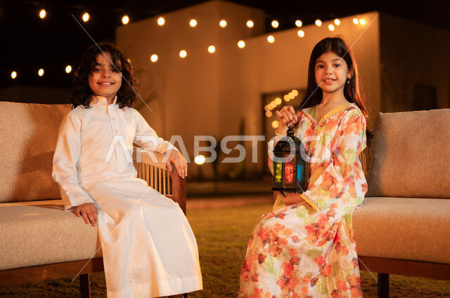 إضاء وزينة الأعياد والمناسبات في المملكة العربية السعودية, بنت عربية خليجية سعودية مبتسمة ترتدي فستان تحمل بيدها فانوس معدني مضيء تجلس برفقه اخيها في حديقة الشاليه، مفهوم المحبة والترابط الاسري ، الاستعداد لاستقبال شهر الخير، العادات والتقاليد السعيدة في شهر رمضان المبارك