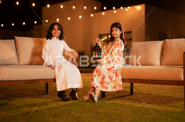 الاستعداد لاستقبال شهر الخير، العادات والتقاليد السعيدة في شهر رمضان المبارك، إضاء وزينة الأعياد والمناسبات في المملكة العربية السعودية, بنت عربية خليجية سعودية مبتسمة ترتدي فستان تحمل بيدها فانوس معدني مضيء تجلس برفقه اخيها في حديقة الشاليه، مفهوم المحبة والترابط الاسري 