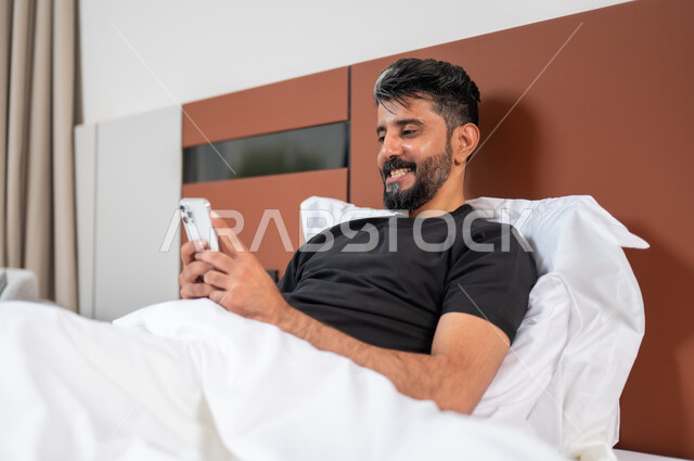 الدردشة والتواصل مع الاصدقاء بإيماءات السعادة والفرح، استخدام الأجهزة والتقنيات الحديثة، صورة مقربة لشاب عربي خليجي سعودي مبتسم يستلقي على السرير يمسك بيده الهاتف المحمول يقوم بتصفح الانترنت ومواقع التواصل الاجتماعي