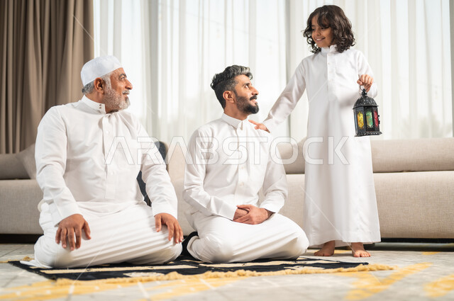 فتى يحمل بيده فانوس معدني مضيء، الإكثار من الطاعة والتقرب الى الله في شهر رمضان المبارك، رجل عربي سعودي خليجي يجلس على سجادة الصلاة في غرفة المعيشة يؤدي فرض الصلاة بجانب ابنه الشاب، الاجتهاد في العبادة واداء الفرائض الدينية، اجواء عائلية رمضانية روحانية