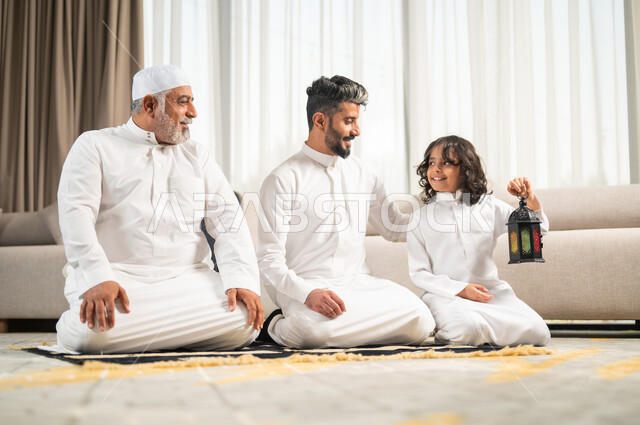 فتى يحمل بيده فانوس معدني مضيء، الإكثار من الطاعة والتقرب الى الله في شهر رمضان المبارك، رجل عربي سعودي خليجي يجلس على سجادة الصلاة في غرفة المعيشة يؤدي فرض الصلاة بجانب ابنه الشاب، الاجتهاد في العبادة واداء الفرائض الدينية، اجواء عائلية رمضانية روحانية