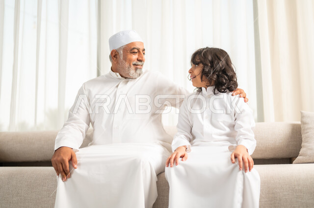 التسامر وتبادل الاحاديث، جلسات عائلية سعيدة، تعابير المودة والمحبة، الاستمتاع بقضاء وقت مع الابناء، صورة مقربة لرجل كبير بالسن عربي خليجي سعودي يرتدي الطاقية البيضاء والثوب التقليدي يجلس على الاريكة بجانب حفيده ي غرفة الجلوس