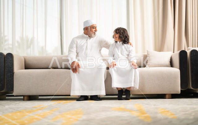 Happy family sessions, expressions of affection and love, enjoying spending time with the children, an elderly Saudi Arabian Gulf man wearing a white hat and traditional dress sitting on the sofa next to his grandson in the living room, chatting and exchanging conversations.