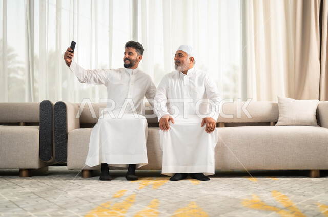توثيق اللحظات الجميلة باستخدام الهاتف المحمول، رجل كبير بالسن عربي خليجي سعودي يرتدي الطاقية البيضاء والثوب التقليدي يجلس على الاريكة بجانب ابنه الشاب في غرفة الجلوس، التسامر وتبادل الاحاديث، جلسات عائلية سعيدة، تعابير المودة والمحبة، التقاط صور سيلفي تذكارية