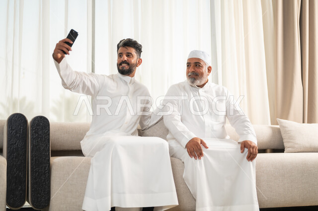 التقاط صور سيلفي تذكارية، توثيق اللحظات الجميلة باستخدام الهاتف المحمول، رجل كبير بالسن عربي خليجي سعودي يرتدي الطاقية البيضاء والثوب التقليدي يجلس على الاريكة بجانب ابنه الشاب في غرفة الجلوس، التسامر وتبادل الاحاديث، جلسات عائلية سعيدة، تعابير المودة والمحبة