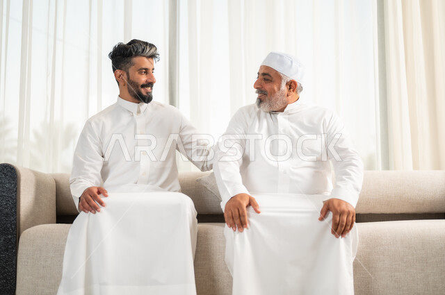 الاستمتاع بقضاء وقت مع الابناء، صورة مقربة لرجل كبير بالسن عربي خليجي سعودي يرتدي الطاقية البيضاء والثوب التقليدي يجلس على الاريكة بجانب ابنه الشاب في غرفة الجلوس، التسامر وتبادل الاحاديث، جلسات عائلية سعيدة، تعابير المودة والمحبة